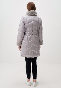 Giacca puffy grigia chiaro con cuciture a zig-zag, colletto e polsini foderati di pelliccia, vita elastica e design lungo fino al ginocchio, abbinata a leggings neri.