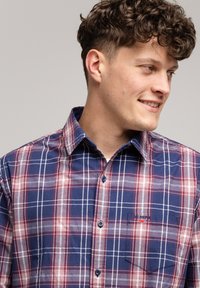 Młody mężczyzna o kręconych włosach, ubrany w niebiesko-czerwoną koszulę w kratę z guzikami, uśmiechający się i patrzący w prawo na jednolitym tle.