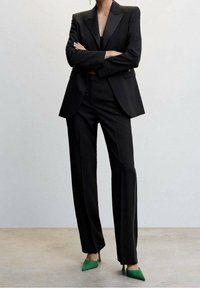 Musta räätälöity puku, jossa on jämäkkä bleiseri, leveälahkeiset housut ja vihreät tikkukorkokengät. Sileä kangas ja terävät kaulukset.