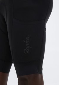 Közelkép fekete kerékpáros nadrágról, oldalzsebbel és diszkrét "Rapha" logóval a combon.