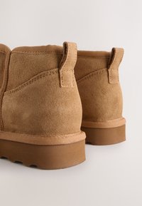 Paire de bottines en daim beige avec languettes et semelles en caoutchouc marron, vues de dos sur un fond uni clair.