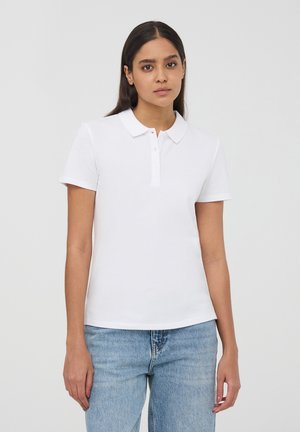 Polo shirt bianco a maniche corte con colletto classico, patta con tre bottoni e tessuto di cotone morbido e liscio. Indossato con jeans azzurri.