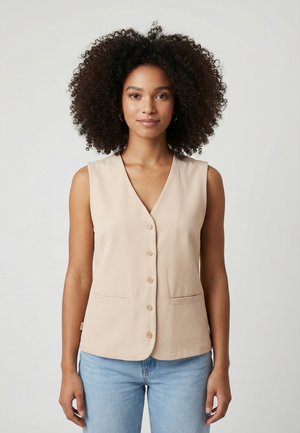 Donna con capelli ricci che indossa un gilet beige senza maniche con bottoni e jeans azzurri, in piedi davanti a uno sfondo semplice.