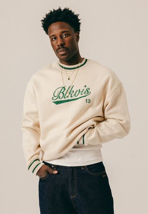 Hombre con cabello corto y rizado, vistiendo una sudadera crema con texto verde "Bkvis 13" y jeans oscuros, de pie con una mano en el bolsillo.