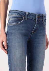 Jeans de mezclilla en azul medio con un corte ajustado. Entre sus características se incluyen bolsillos frontales, un cierre de botón y detalles de costura visibles a lo largo de las costuras.
