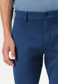 Pantalon bleu marine avec une finition lisse et texturée. Présente une fermeture à bouton et des passants de ceinture, avec un gros plan sur la taille et la main.