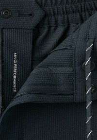 Gros plan d'une veste noire texturée avec fermeture éclair, bouton, cordon et texte visible « MNG PERFORMANCE » sur le tissu intérieur.