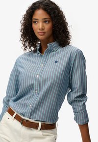 Chemise à col boutonné à rayures bleues et vertes, en tissu de coton, avec un petit logo sur la poitrine, assortie à une ceinture marron et un pantalon clair.