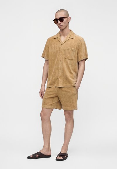 Homme portant une chemise à manches courtes couleur sable avec boutons et un short assorti, des lunettes de soleil foncées et des sandales à enfiler foncées, debout devant un fond uni.