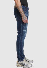 Vaqueros ajustados en azul oscuro con áreas desgastadas y parches desgastados. Combinados con zapatillas deportivas en plata y oro.