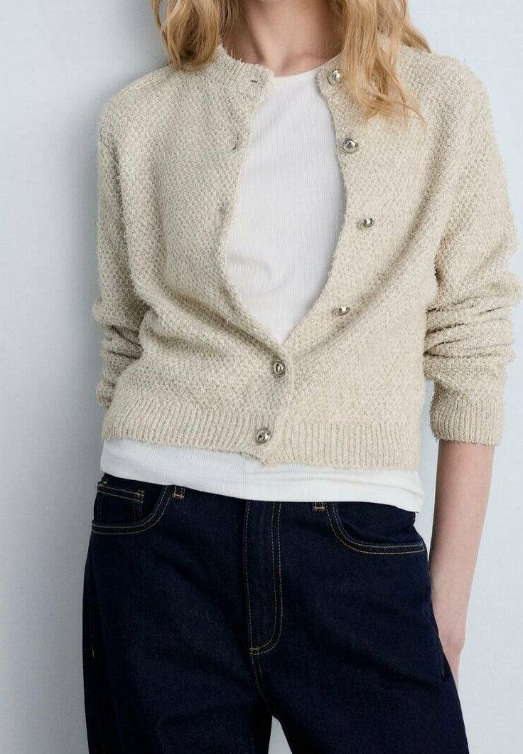 Femme portant un cardigan beige texturé boutonné en bas par-dessus une chemise blanche, associé à un jean bleu foncé, debout les mains dans les poches.