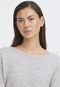 Maglione grigio dalla vestibilità ampia, con scollo rotondo e perle decorative sparse sul tessuto. Texture morbida, maniche lunghe, design minimalista.