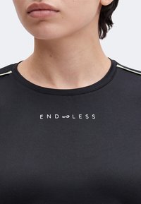 Gros plan d'une personne portant un t-shirt noir avec le mot "ENDLESS", où les doubles O sont remplacés par un symbole infini.