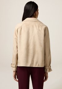 Veste en daim beige avec une coupe décontractée, dotée d'un grand col, de manches raglan et de poignets ceinturés. Texture lisse, design minimaliste, sans accessoires visibles.