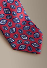 Sarkana zīda kaklasaite ar atkārtotu zilas un baltas ovālas un rombveida formas rakstu ar sarežģītām detaļām. Gluda tekstūra un smaili gals.