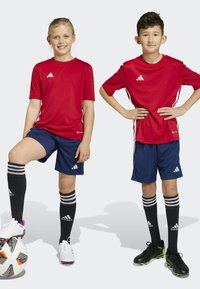 Camisetas deportivas rojas con rayas blancas, pantalones cortos azul marino, calcetas negras con rayas grises y botas de fútbol. Un niño está de pie sobre un balón de fútbol.