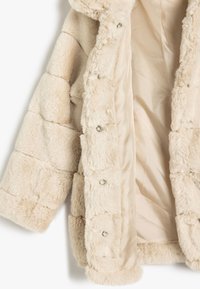 Beige Kunstfelljacke mit einem gesteppten Design, ausgestattet mit glänzenden silbernen Druckknöpfen und einem glatten Satinfutter. Weicher, strukturierter Außenstoff.