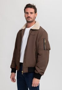 Hombre con chaqueta bomber marrón con cuello de borrego, camisa blanca y vaqueros azules sobre un fondo liso gris claro.