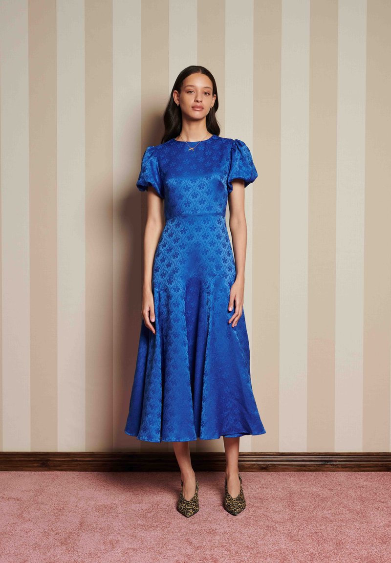 Γυναίκα που στέκεται μπροστά σε καρό τοίχο σε μπεζ αποχρώσεις, φορώντας ένα μπλε midi φόρεμα με μοτίβα και φουσκωτά μανίκια, καθώς και τακούνια με λεοπάρ σχέδιο.