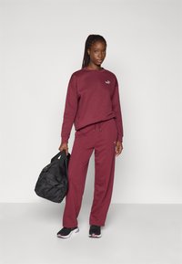Mørkerød sweatshirt og joggers lavet af blødt stof, med et lille logo. Sort duffel taske med håndtag, båret med sorte sneakers.