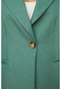 Teal-puku, jossa on viilattava kaulus, yksi suuri ruskea nappi ja etutaskut. Silkkinen kangas, jossa on hienovarainen rakenne.