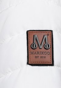 Vit vadderad tyg med en brun läderlogotyp, med präglade bokstäver och en distinkt kant. Texten inkluderar "MARIKOO EST 2016".