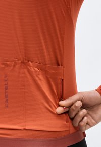 Χέρι που ρυθμίζει το φερμουάρ σε μικρή πίσω τσέπη ενός ποδηλατικού τζέρσεϊ Castelli σε σκουρόχρωμο σκουριά, με υφή υφάσματος.