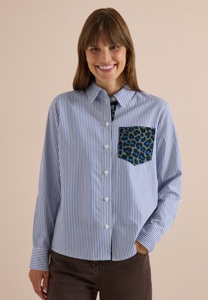 Femme souriante portant une chemise boutonnée à rayures bleues et blanches avec une poche poitrine à imprimé léopard bleu et noir et un pantalon foncé.