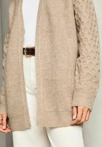 Beige kardigán bordázott textúrával és buborékos kötésű ujjal. Nyitott elülső kialakítás, közepes hosszúságú, krém színű nadrággal és övvel párosítva.