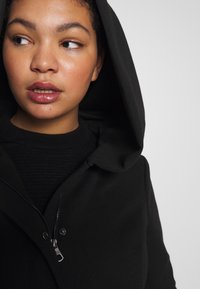 Melna ar rāvējslēdzēju jaka ar lielu, karājošu kapuci, mīkstu tekstūru un redzamu rāvējslēdzēja detaļu. Valkāta pār teksturētu melnu augšu.