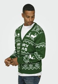 Grüne gestrickte Strickjacke mit weißen, weihnachtlichen Mustern, darunter Rehe und Schneeflocken. Ribbenbesätze an den Ärmeln und ein V-Ausschnitt.