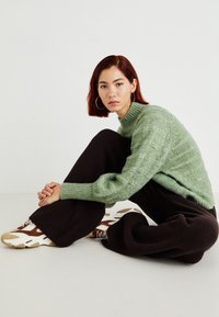 Pull en maille texturée vert avec un col haut, associé à un large pantalon noir côtelé et des baskets blanches avec des accents marron.
