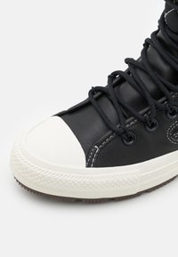 Svart läder hög topp sneakers med en vit gummikappa, runda svarta  öglor och texturerade svarta snörningar, med vita sömnadsdetaljer.