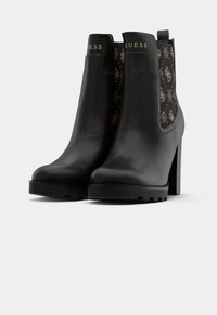 Bottines en cuir noir avec un panneau en tissu noir texturé arborant un logo doré. Talon épais et semelle en caoutchouc.