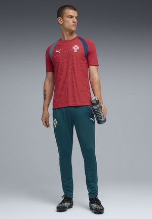 Uomo con divisa da calcio rossa e blu del Portogallo che tiene una bottiglia d'acqua, in piedi davanti a uno sfondo grigio uniforme.