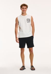 Weißes ärmelloses Baumwolltop mit einem schwarzen grafischen Design auf der Brust, kombiniert mit schwarzen Shorts und schwarzen Schlippers.