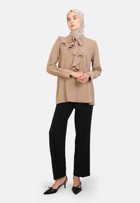 Beige blouse met ruffles bij de halslijn, lange mouwen en zwarte broek gecombineerd met puntige zwarte hakken. Glad materiaal, getailleerde pasvorm.