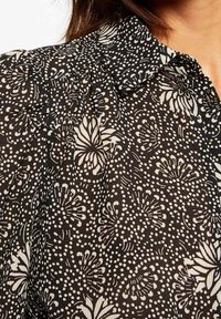 Blouse en tissu noir avec un motif floral et à pois crème. Le design comprend un col classique et des détails froncés aux épaules.