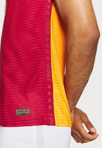 Jersey deportivo de Nike con franjas verticales rojas y naranjas, tejido texturizado y una etiqueta dorada. Texto "1905" en color marrón a lo largo de la costura lateral.