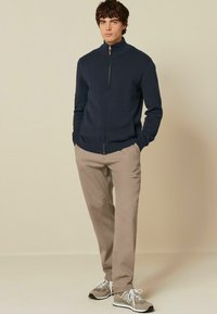 Marineblauer Reißverschluss-Pullover mit hohem Kragen, kombiniert mit beigen Hosen und grauen Sneakers mit weißen Akzenten. Glatte Textur, lässige Passform.