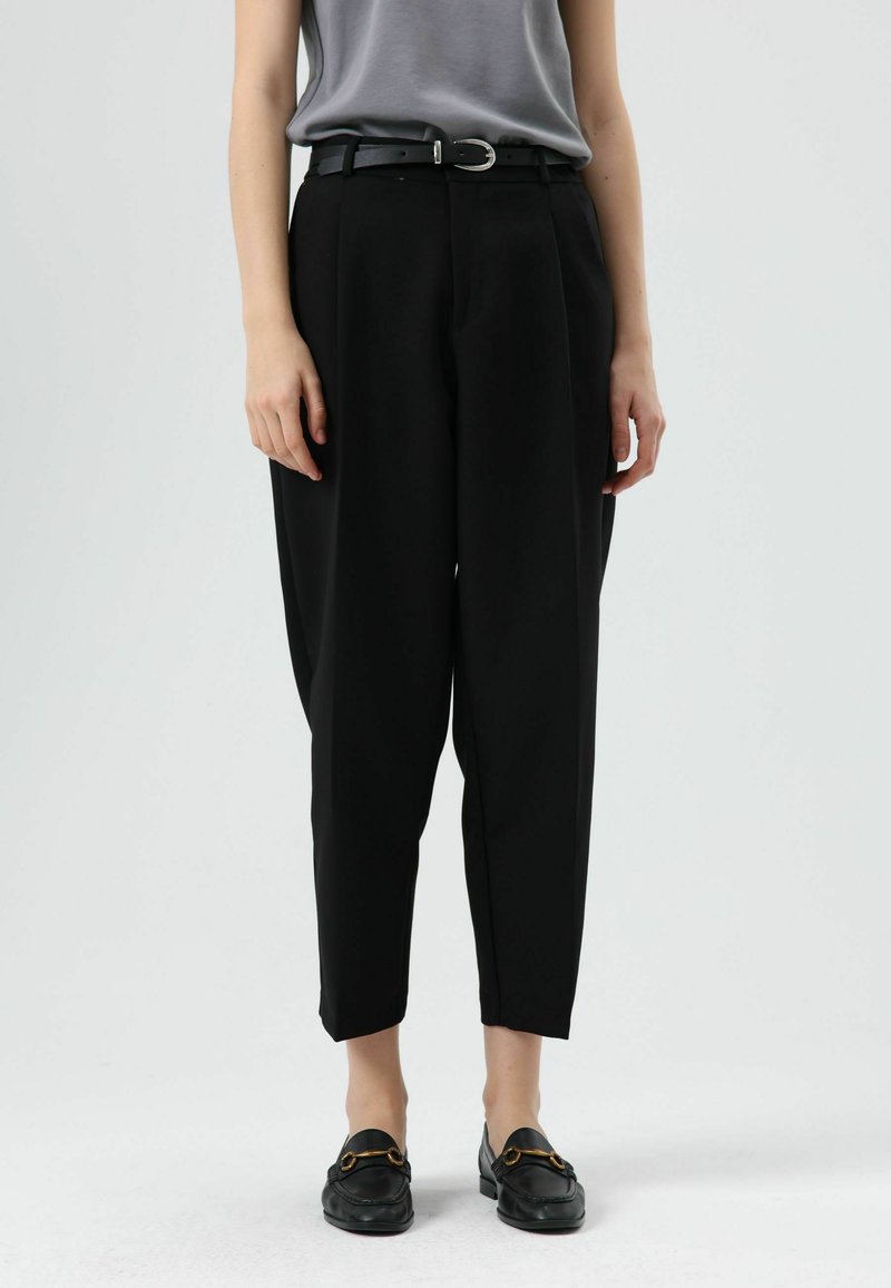 Pantalon noir fuselé taille haute, doté d'une ceinture simple avec une boucle argentée. Porté avec des mocassins noirs plats. Fini en tissu lisse.