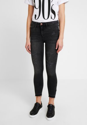 Personne portant un jean skinny noir déchiré avec des ourlets bruts, un t-shirt graphique blanc des années 90, et des baskets noires avec des semelles blanches.