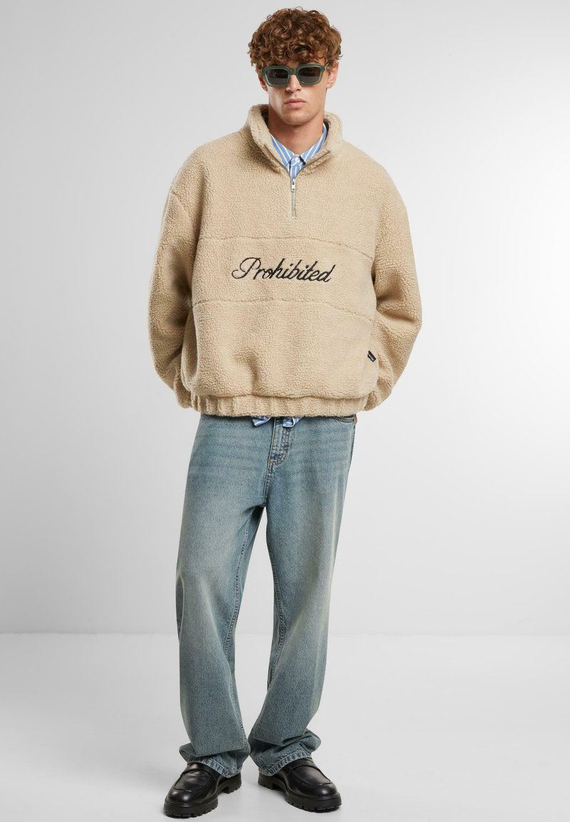 Pullover in pile beige con colletto a mezzo zip e ricamo nero "Proibito" sul petto, abbinato a jeans azzurri chiari e scarpe nere.
