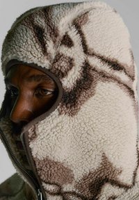 Fleecehættetrøje med lynlåslukning, med en cremefarvet base og brune abstrakte mønstre. Blød tekstur og tætsiddende pasform omkring ansigtet.