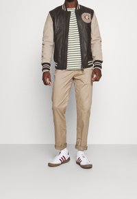 Man i brun och beige varsityjacka, grön och vit randig skjorta, beige upprullade byxor, vita sneakers med röda ränder, stående.