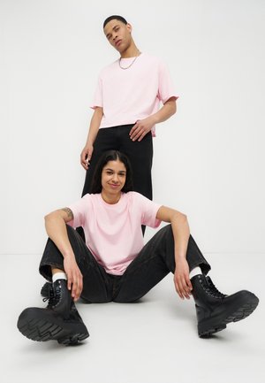 Deux jeunes adultes portent des t-shirts rose clair et un pantalon noir ; l'un est assis par terre en souriant, l'autre se tient debout derrière avec une expression neutre.
