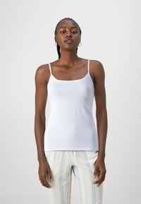 Top tanque branco sem mangas com um decote quadrado, feito de tecido suave. Modelo veste calças listradas com tons de azul e creme.