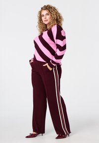 Fuchsia en donkerpaarse gestreepte gebreide trui gecombineerd met bordeauxrode broek met witte zijstrepen; glanzende bordeauxrode hakken maken de look compleet.