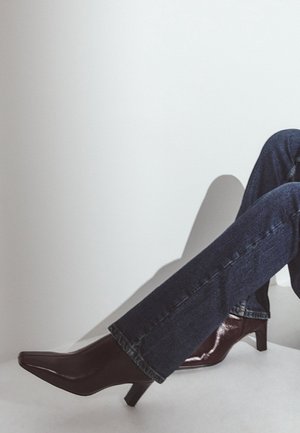 Person trägt dunkelblaue Jeans und dunkelbraune Leder-Stiefeletten mit Absatz und sitzt vor einem schlichten hellen Hintergrund.