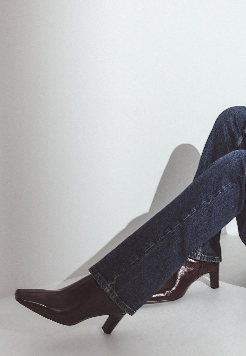 Personne portant un jean bleu foncé et des bottines à talons en cuir marron foncé assise devant un fond clair uni.